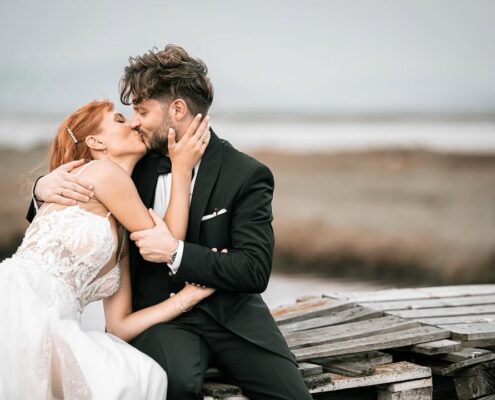 Ο γαμπρός και η νύφη κάθονται σε μια ξύλινη προβλήτα δίπλα στο νερό.