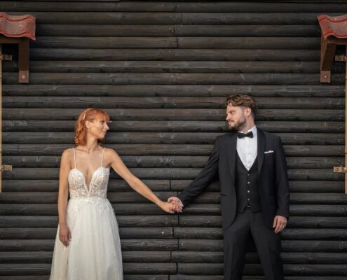 Το ζευγάρι στέκεται κρατώντας τα χέρια με φόντο έναν μοντέρνο σκούρο ξύλινο τοίχο.