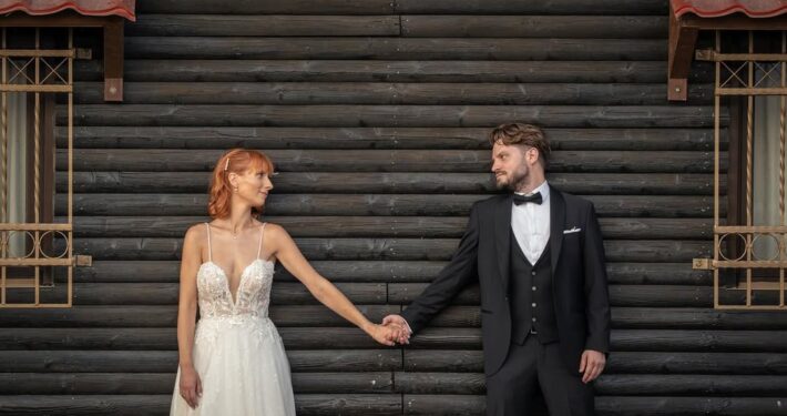 Το ζευγάρι στέκεται κρατώντας τα χέρια με φόντο έναν μοντέρνο σκούρο ξύλινο τοίχο.