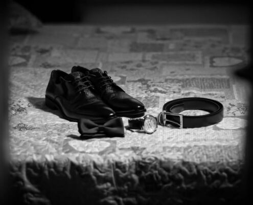 Flatlay ασπρόμαυρη λήψη με τα παπούτσια, το παπιγιόν, τη ζώνη και το ρολόι του γαμπρού.
