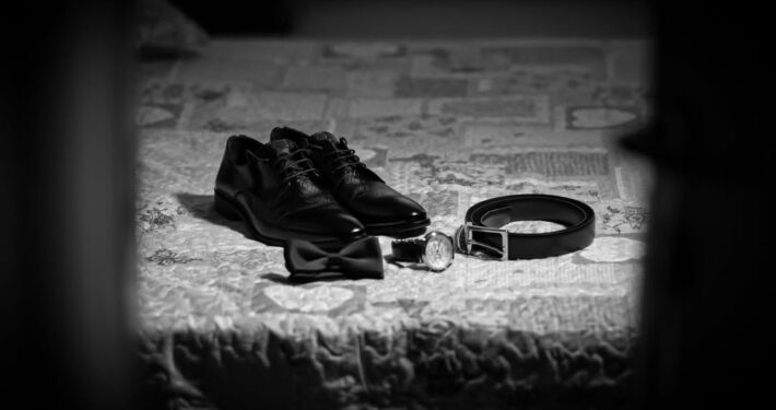 Flatlay ασπρόμαυρη λήψη με τα παπούτσια, το παπιγιόν, τη ζώνη και το ρολόι του γαμπρού.