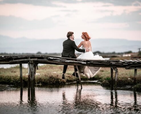 Ο γαμπρός και η νύφη κάθονται αγκαλιασμένοι σε μια μικρή ξύλινη γέφυρα πάνω από το νερό.