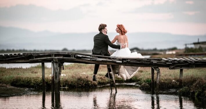 Ο γαμπρός και η νύφη κάθονται αγκαλιασμένοι σε μια μικρή ξύλινη γέφυρα πάνω από το νερό.