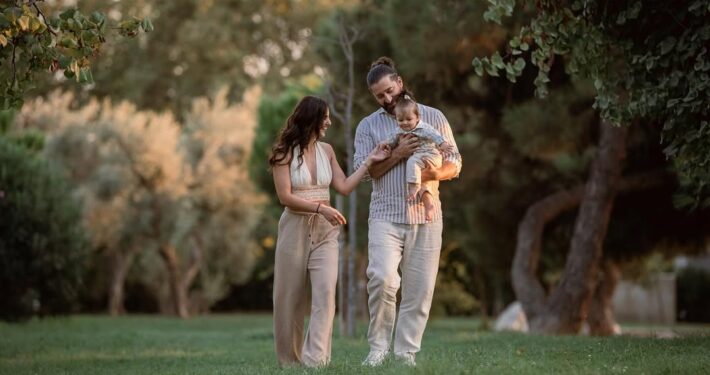 Οικογένεια περπατάει ανέμελα στο πάρκο, με τον πατέρα να κρατάει το μωρό αγκαλιά.