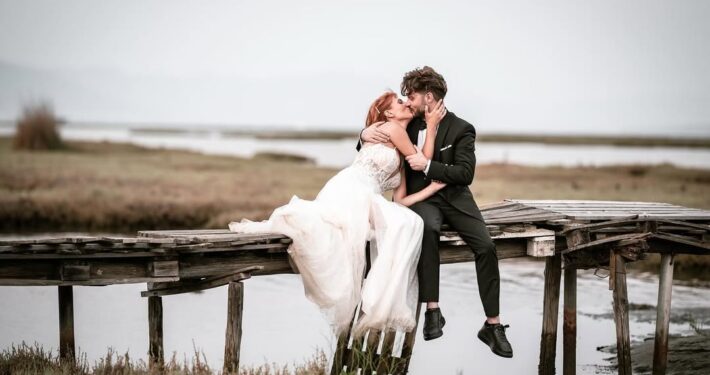 Το ζευγάρι κάθεται σε μια ξύλινη αποβάθρα δίπλα στο νερό και ανταλλάσσει ένα φιλί.