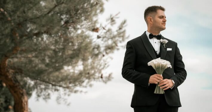 Ο γαμπρός με μαύρο κοστούμι κρατάει τη νυφική ανθοδέσμη σε εξωτερικό χώρο.