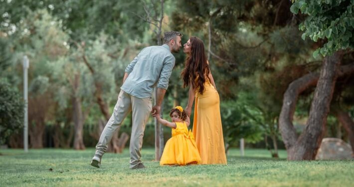 Οικογένεια περπατάει στο γρασίδι, με τους γονείς να ανταλλάσσουν ένα φιλί και το παιδί να προπορεύεται.