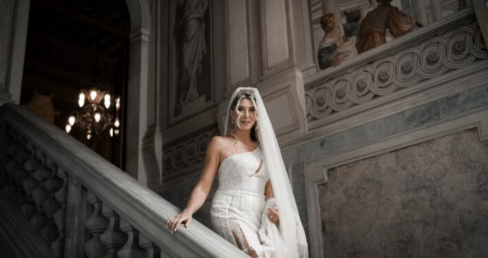 Η νύφη με το νυφικό της στέκεται στα σκαλιά μιας μεγαλοπρεπούς μαρμάρινης σκάλας.