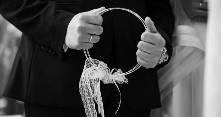 Κοντινή ασπρόμαυρη λήψη στα χέρια του γαμπρού που κρατούν τα γαμήλια στέφανα.