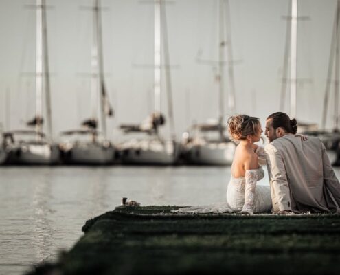 Το νιόπαντρο ζευγάρι κάθεται σε μια ξύλινη αποβάθρα με ιστιοπλοϊκά σκάφη στο φόντο.