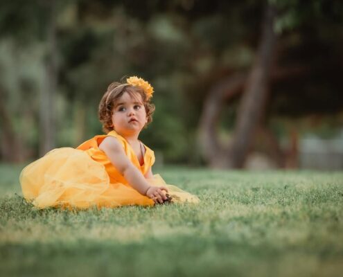 Ένα μικρό κορίτσι με κίτρινο φόρεμα κάθεται στο γρασίδι και κοιτάζει τον φακό.