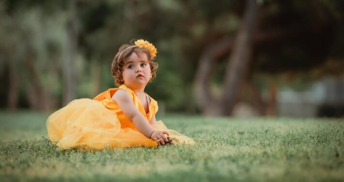 Ένα μικρό κορίτσι με κίτρινο φόρεμα κάθεται στο γρασίδι και κοιτάζει τον φακό.