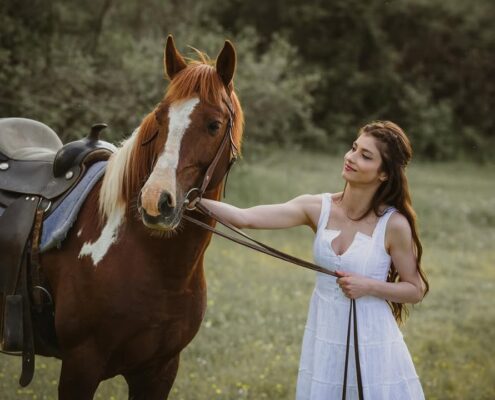 Η νύφη με λευκό φόρεμα χαμογελάει κρατώντας τα ηνία ενός καφέ αλόγου στη φύση.