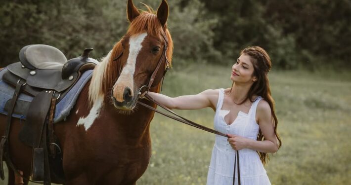 Η νύφη με λευκό φόρεμα χαμογελάει κρατώντας τα ηνία ενός καφέ αλόγου στη φύση.