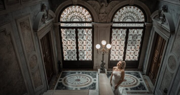 Γάμος στην Βενετία. Η νύφη ποζάρει δίπλα σε μια μεγάλη περίτεχνη πόρτα με βιτρό σε ένα εντυπωσιακό κτίριο.